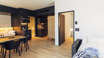 Una habitación de hotel moderna con suelo de madera, zona de comedor con mesa y sillas, kitchenette y cama con manta y un peluche.