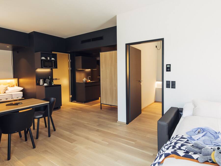 Una habitación de hotel moderna con suelo de madera, zona de comedor con mesa y sillas, kitchenette y cama con manta y un peluche.