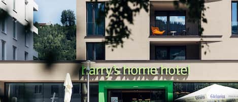 A hotel named "Harry's Home Hotel" with a green entrance, outdoor seating, and a view of a hill.
