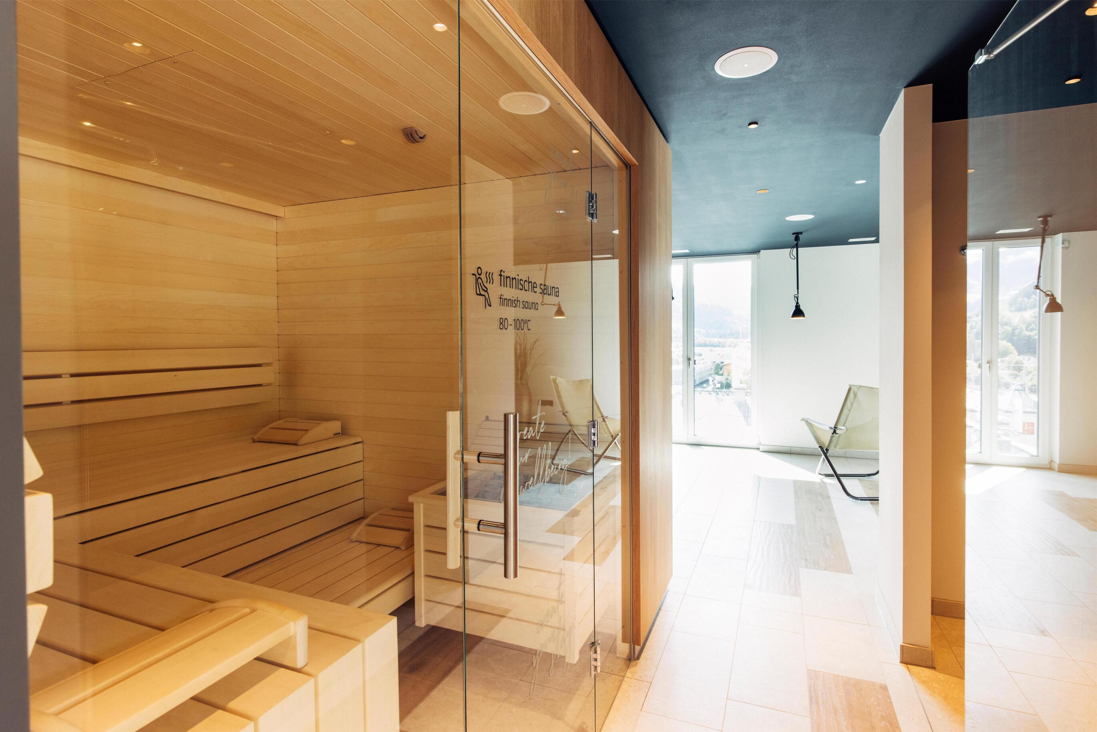A modern sauna with wooden benches and glass doors, a seating area with chairs, and a view of the outdoors through large windows.