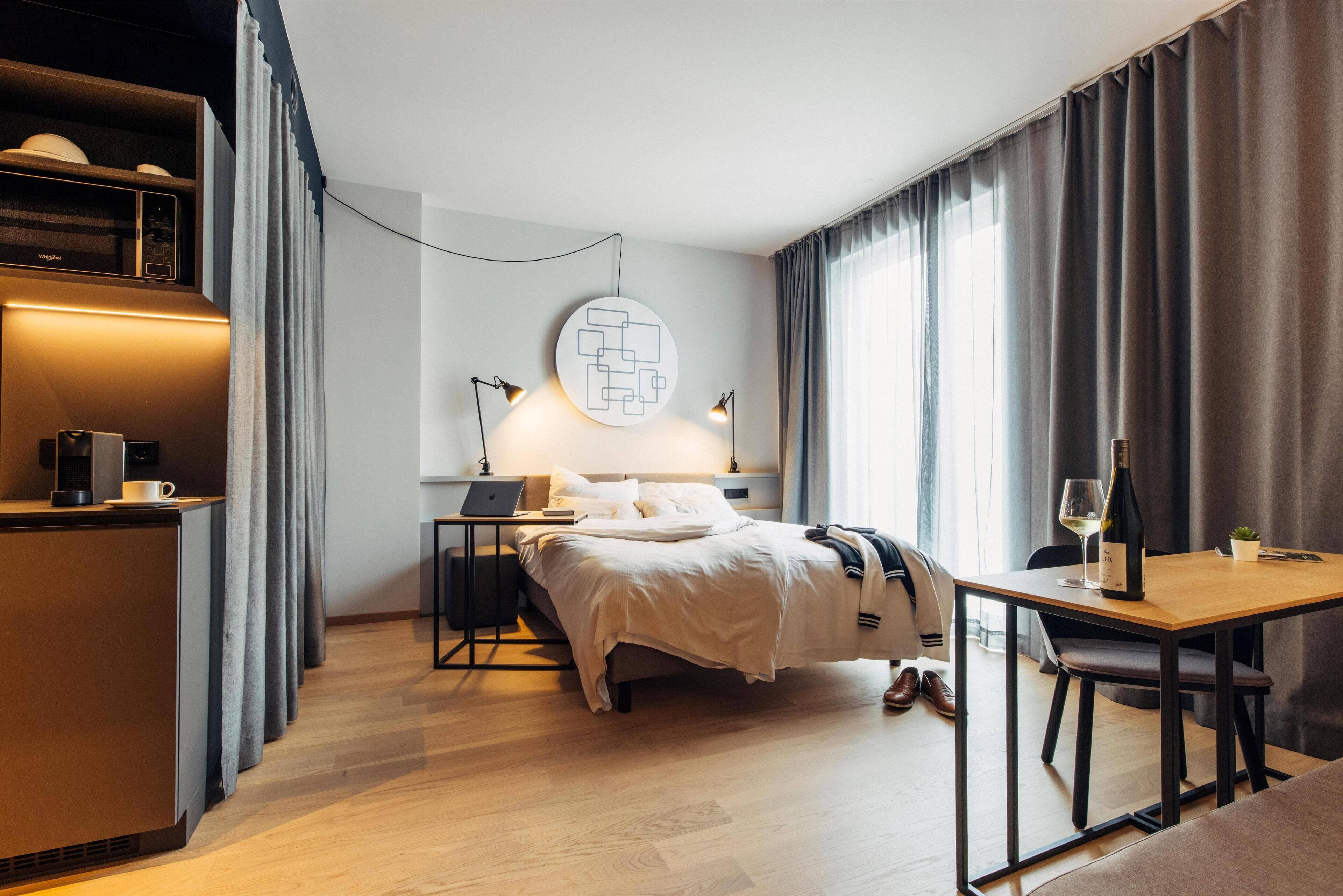 A modern hotel room with a bed, a desk, a laptop, and a wine bottle.