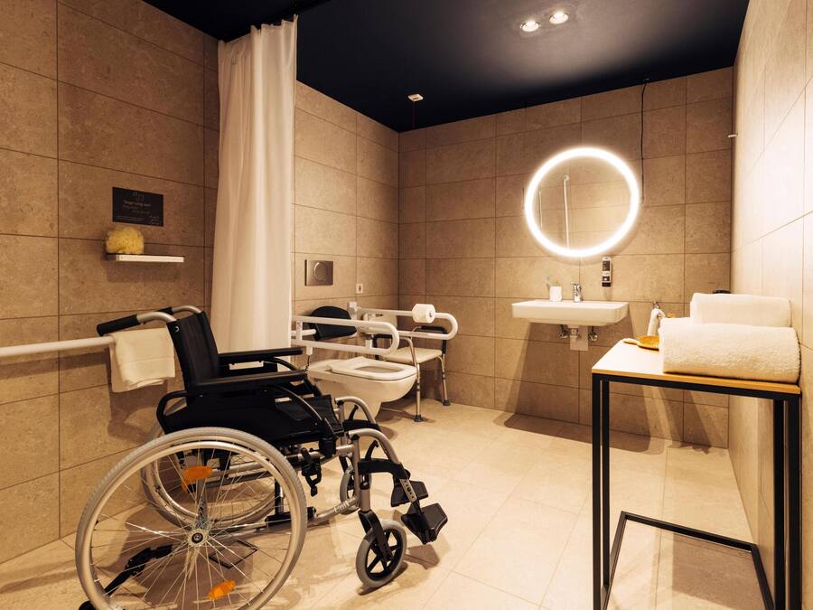 A modern bathroom with a wheelchair, a toilet, a sink, and a mirror with a circular light.