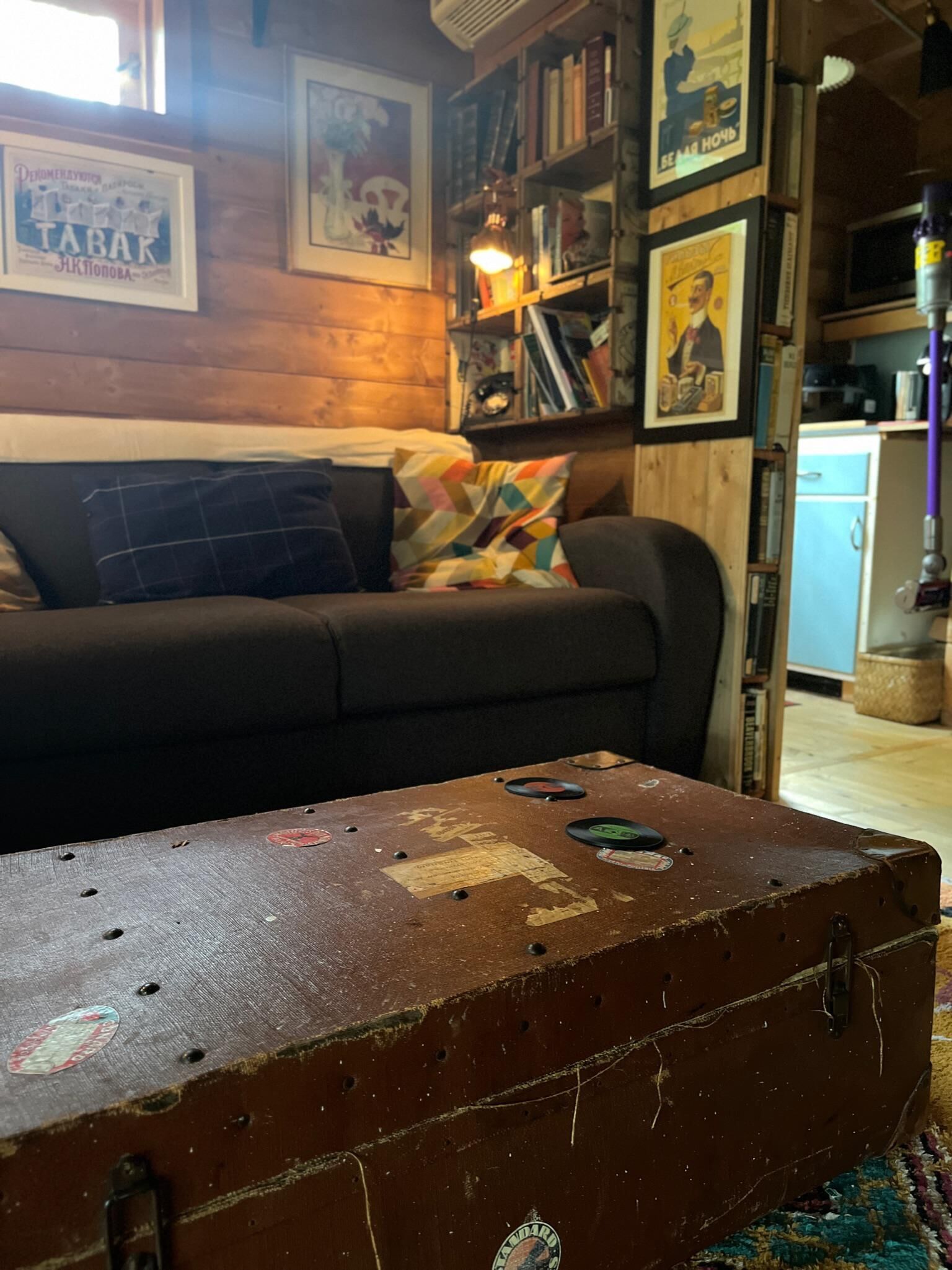 Ein gemütliches Wohnzimmer mit einem Vintage-Koffer auf einem Teppich, einer dunklen Couch mit bunten Kissen und einem Bücherregal voller Bücher und gerahmter Bilder.
