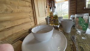 Un intérieur en bois avec une table dressée pour le repas, comprenant un bol blanc, une tasse blanche et un verre orné d’un motif végétal.