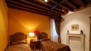 A room with two beds, a small table with a lamp, a vase with flowers, a fireplace, and a painting on the wall.