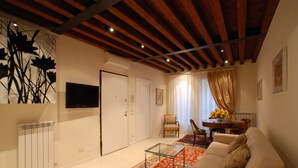 A living room with a sofa, a glass coffee table, a patterned rug, a wooden ceiling, and a TV mounted on the wall.