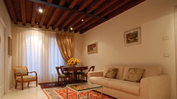 A living room with a sofa, armchairs, a glass coffee table, a red patterned rug, wooden ceiling beams, and two framed pictures on the wall.