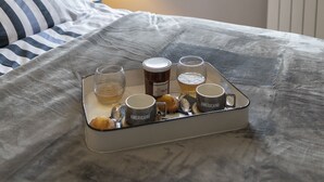 A neatly made bed with a tray of breakfast items including coffee, tea, and pastries, a bedside lamp, and a window with a view of greenery.