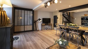 A modern living room with a wooden floor, a dining area with metal chairs, a TV, and a staircase leading to an upper level.
