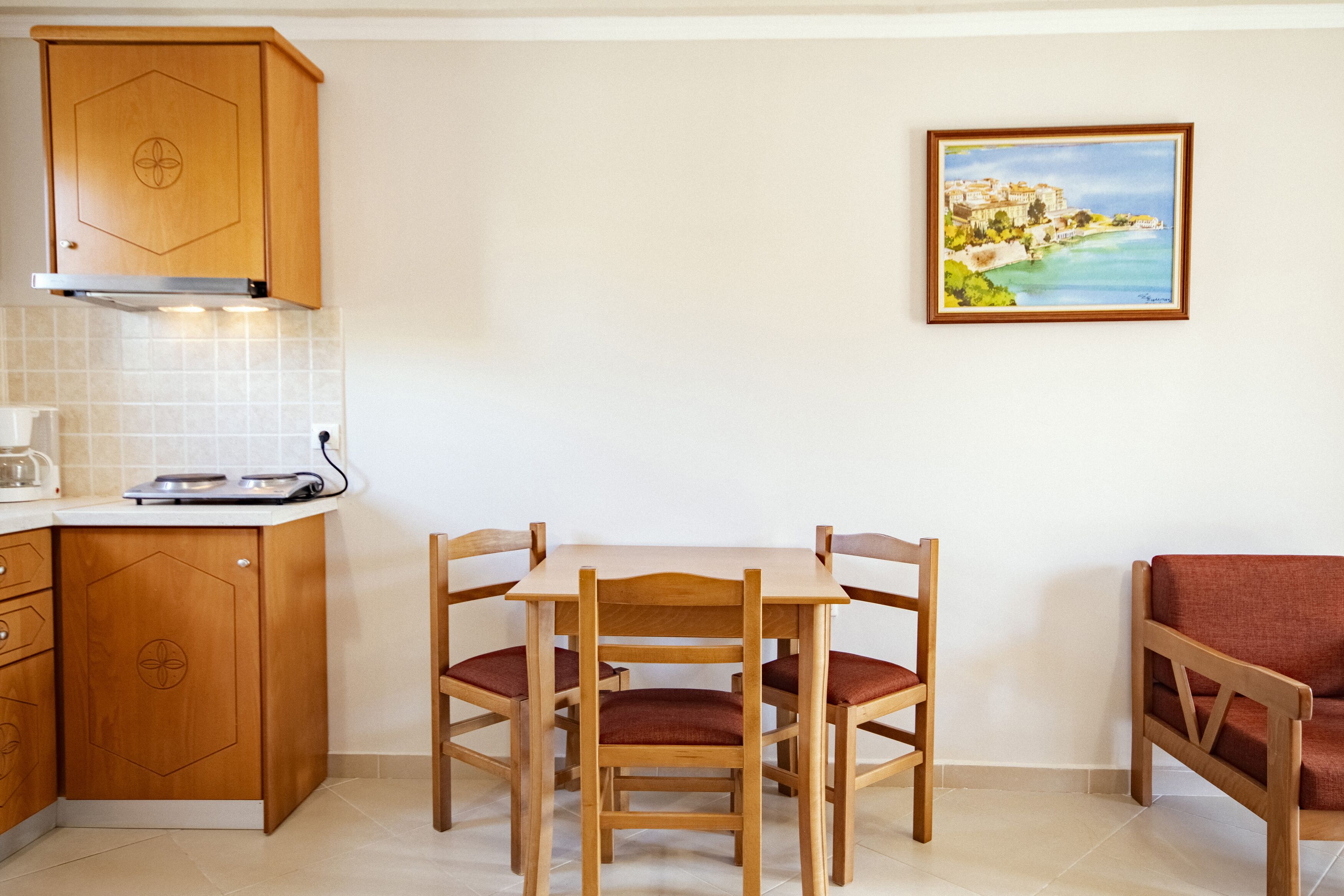 A compact kitchen with wooden cabinets, a dining table with chairs, and a painting of a coastal scene on the wall.