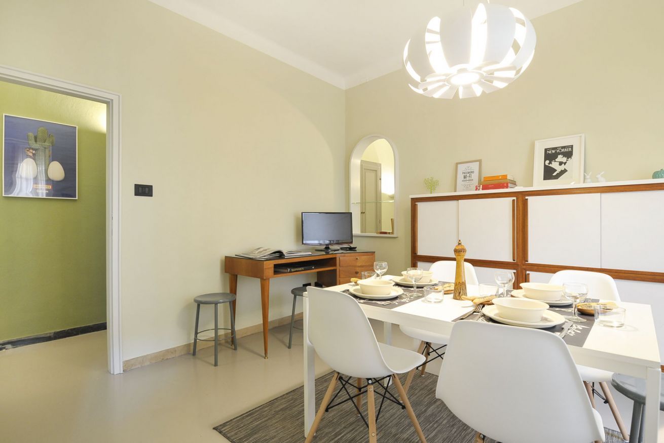 Uma área de refeições com uma mesa branca posta para quatro pessoas, uma televisão sobre uma escrivaninha de madeira e um espelho acima da mesa.