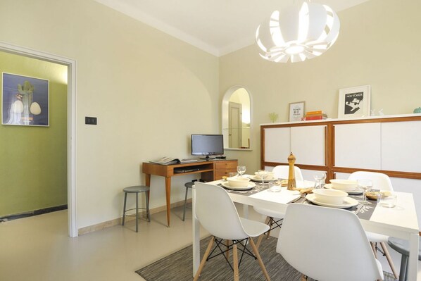 Un comedor con una mesa blanca dispuesta para cuatro, un televisor sobre un escritorio de madera y un espejo sobre la mesa.