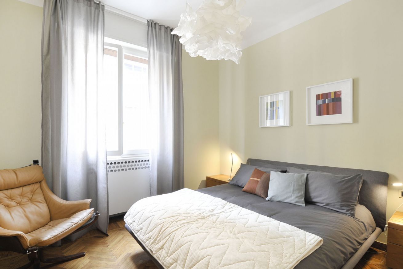 A bedroom with a bed, a chair, a window with curtains, and two framed pictures on the wall.