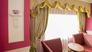 A room with a pink wall, a window with yellow and beige curtains, a white shelf with a decorative item, and a red patterned chair with a round table. - Tokyo Bay Maihama Hotel First Resort (Urayasu)