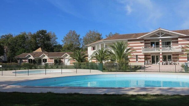 A large building with a swimming pool, surrounded by greenery and a clear blue sky.