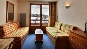 A cozy room with a sofa, a small table, and a wooden dresser. A window offers a view of a snowy landscape. - Residence Le Cervin (La Plagne-Tarentaise)