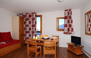 Une pièce chaleureuse avec une table à manger en bois, deux chaises, un canapé rouge, un téléviseur posé sur un meuble en bois et une fenêtre avec des rideaux.