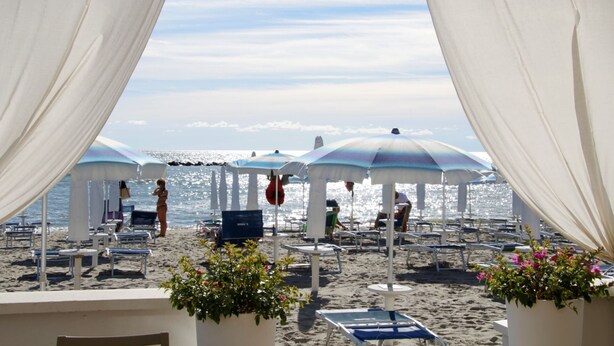 En strand med vita parasoller och solstolar, en klar himmel och havet i bakgrunden.