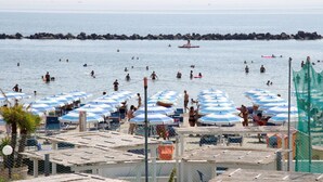 En strand med flera parasoller och solstolar, en stenmur som vågbrytare i bakgrunden och folk som badar i vattnet.