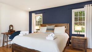 Ein Schlafzimmer mit einem großen Bett, Nachttischen, einem Schreibtisch und zwei mit Vorhängen versehenen Fenstern.