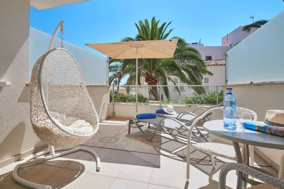 A balcony with a hanging chair, a table with chairs, a sun umbrella, and a bottle of water.