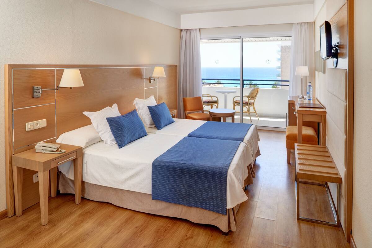A hotel room with a large bed, a desk, a chair, and a balcony with a view of the sea.