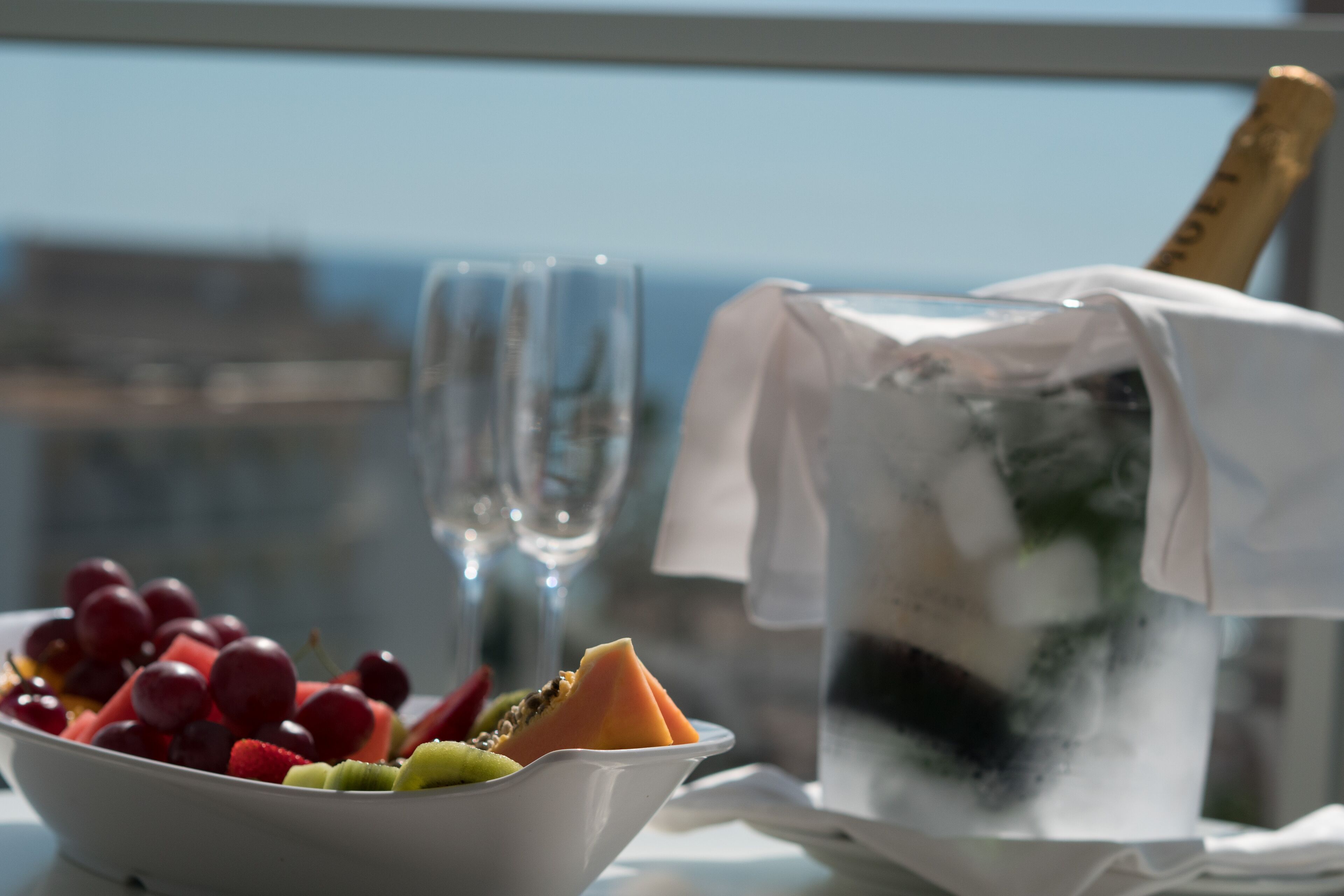 En skål med frukt, ett glas vatten och en flaska champagne står på ett bord med utsikt över staden.