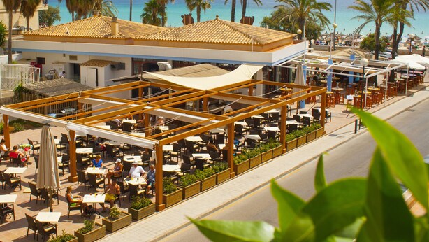 An outdoor café with tables and chairs, overlooking a beach with palm trees and the sea. - Hotel Girasol (Sant Llorenc des Cardassar)