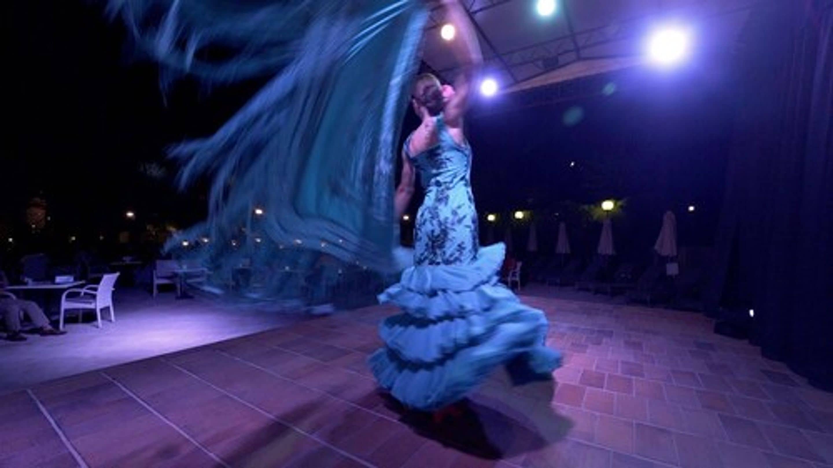 A person in a flowing dress performing on a stage with chairs and tables in the background.