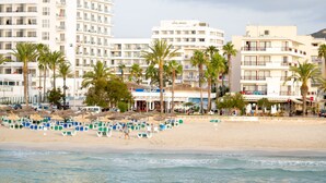 A beach with rows of beach chairs and umbrellas, palm trees, and buildings in the background. - Hotel Girasol (Sant Llorenc des Cardassar)