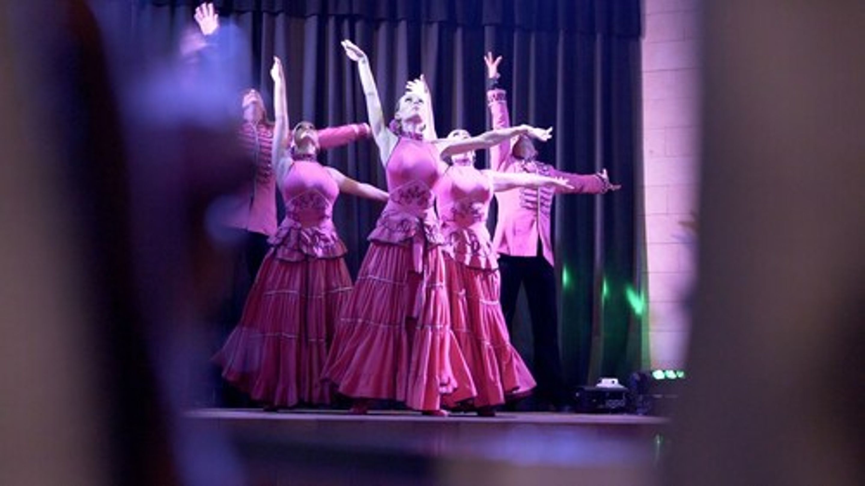 A group of dancers in pink costumes performing on a stage with a dark curtain backdrop.