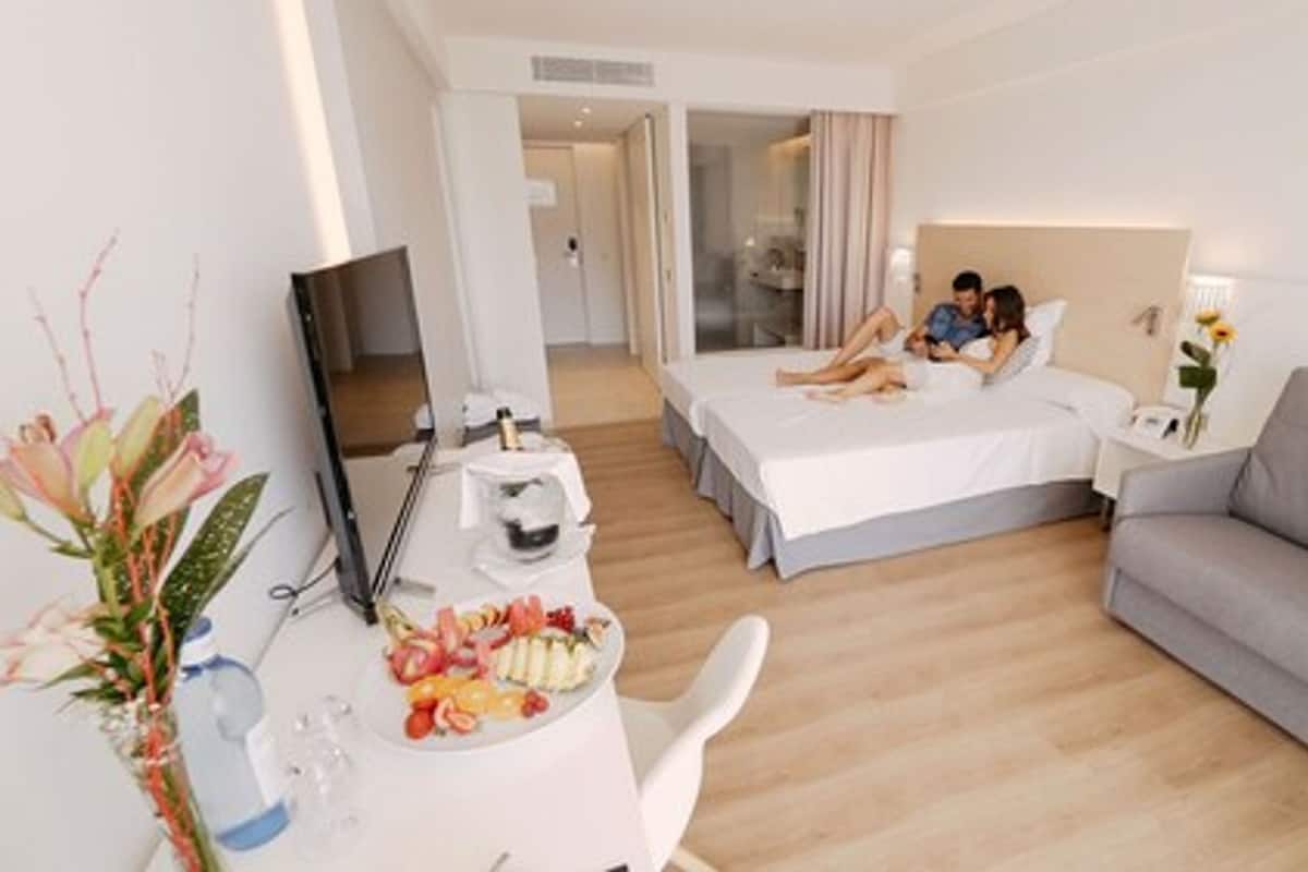 A hotel room with a bed, a sofa, a desk with a laptop, a TV, and a dining table with fruit.