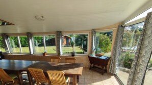 A sunroom with a large table and chairs, large windows with curtains, and a view of a garden. - Logement Locatif à Belle-ile : Maison Avec Jardin Partagé (Le Palais)
