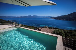 Ein Pool mit klarem, blauem Wasser, umgeben von einer Sonnenterrasse und Blick auf Meer und Berge.