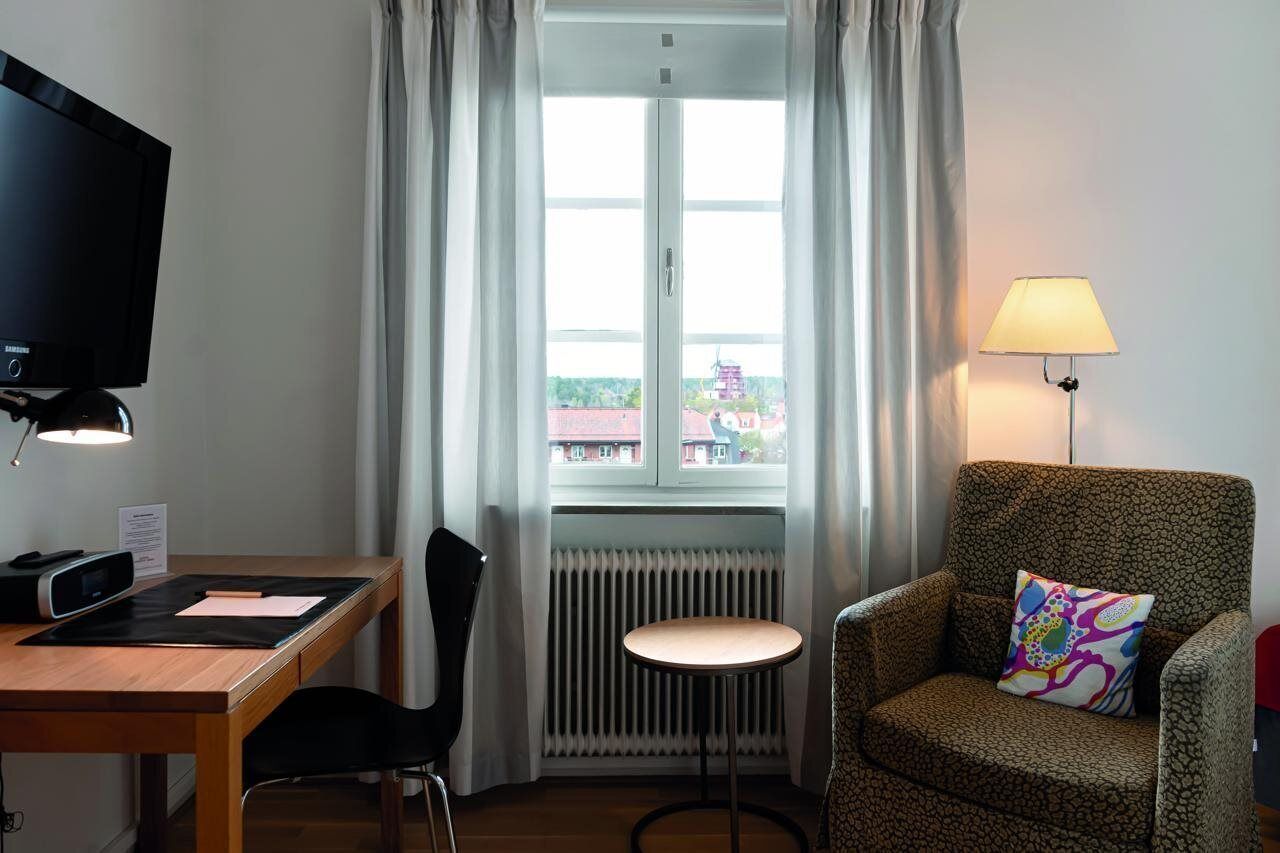 Une chambre d’hôtel équipée d’un bureau, d’une chaise, d’une télévision, d’une fenêtre avec des rideaux, d’un radiateur et d’un fauteuil à motifs agrémenté d’un coussin décoratif.