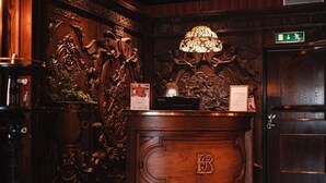 A reception desk with a 'B' monogram, ornate wood carvings, a stained glass lamp, and a checkered floor. - Hotel Bishops Arms Strangnas (Strängnäs)