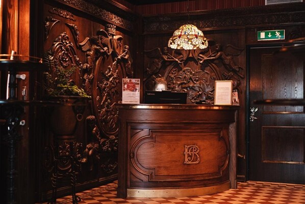 A reception desk with a 'B' monogram, ornate wood carvings, a stained glass lamp, and a checkered floor.