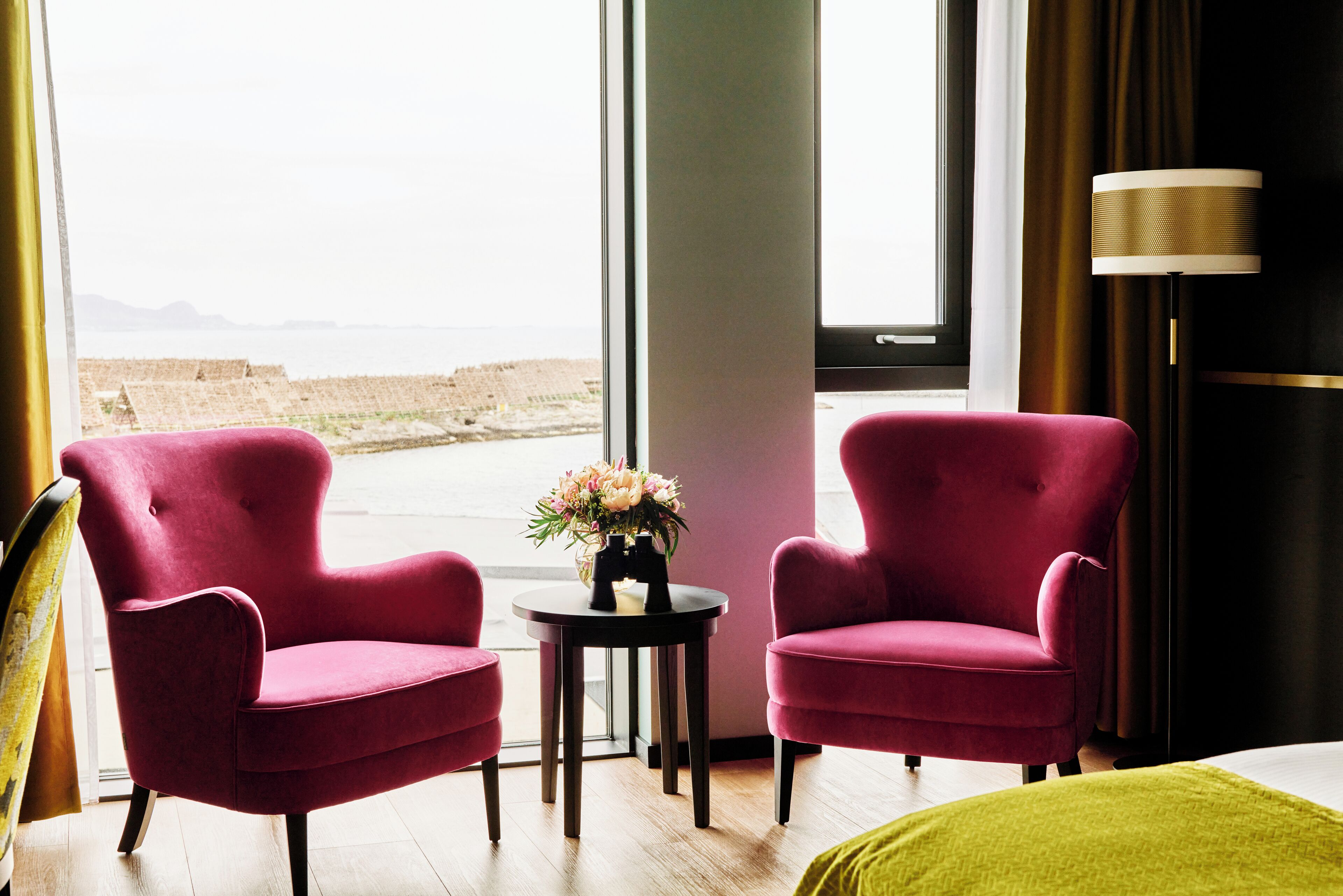 Ein Zimmer mit zwei roten Sesseln, einem kleinen Tisch mit einer Blumenvase und Blick auf den Strand durch ein großes Fenster.
