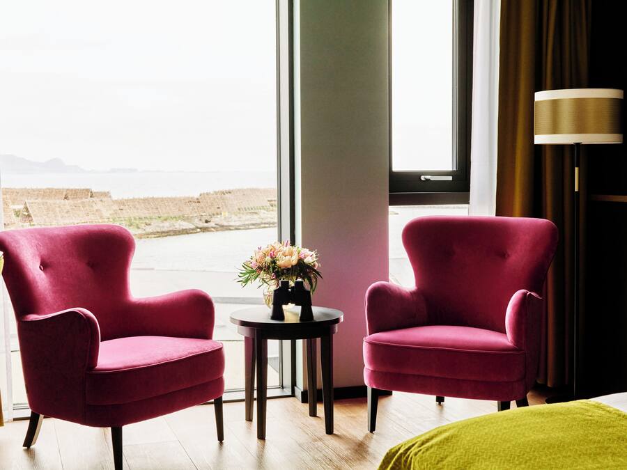 Ein Zimmer mit zwei roten Sesseln, einem kleinen Tisch mit einer Blumenvase und Blick auf den Strand durch ein großes Fenster.