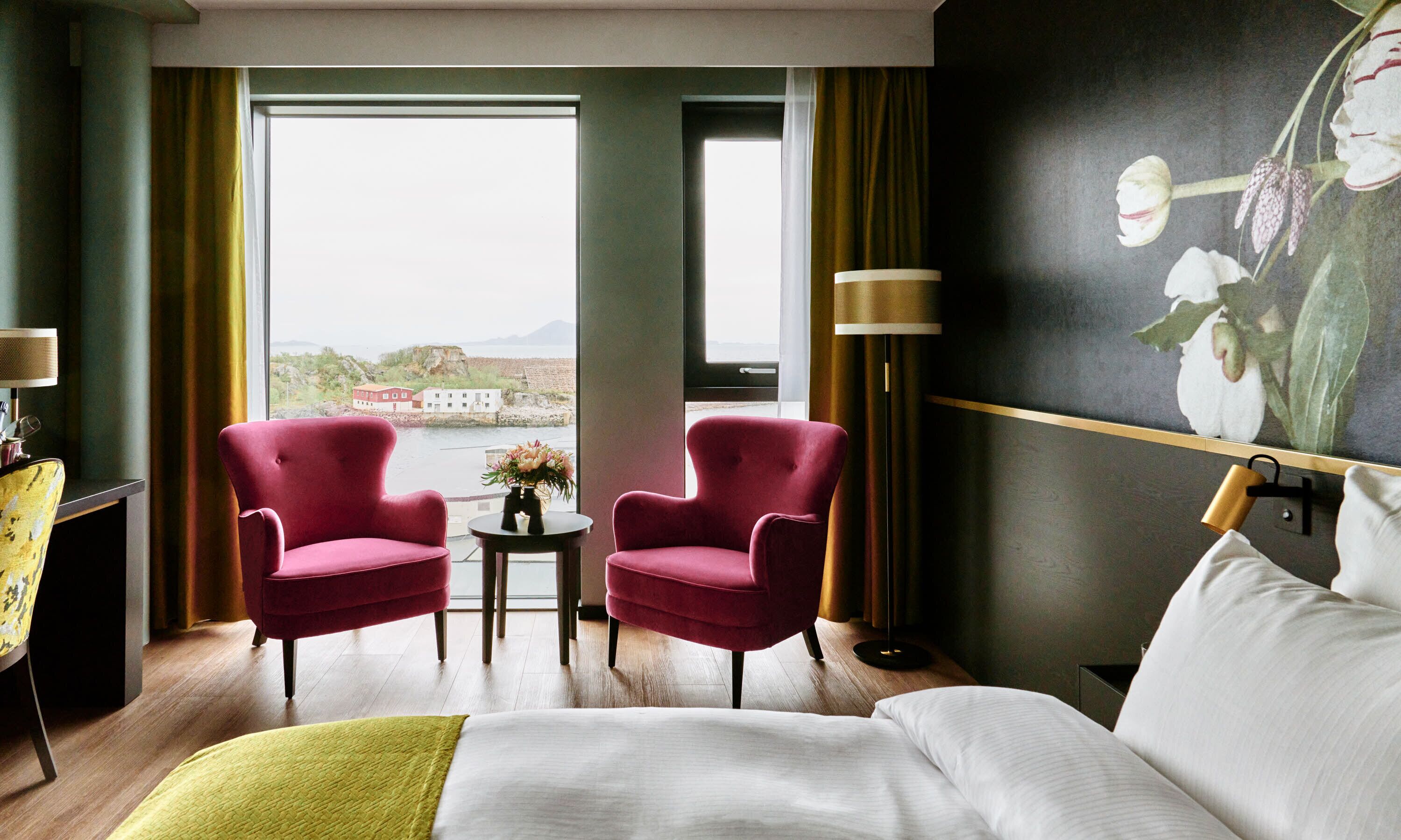 Habitación de hotel con cama, dos sillones rojos, una mesita con un jarrón de flores, una lámpara y un ventanal con vistas a edificios y agua.