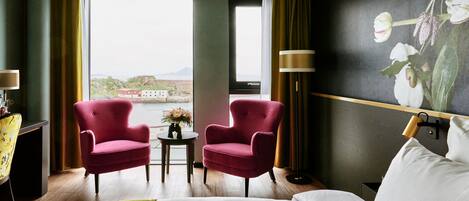 Habitación de hotel con cama, dos sillones rojos, una mesita con un jarrón de flores, una lámpara y un ventanal con vistas a edificios y agua.