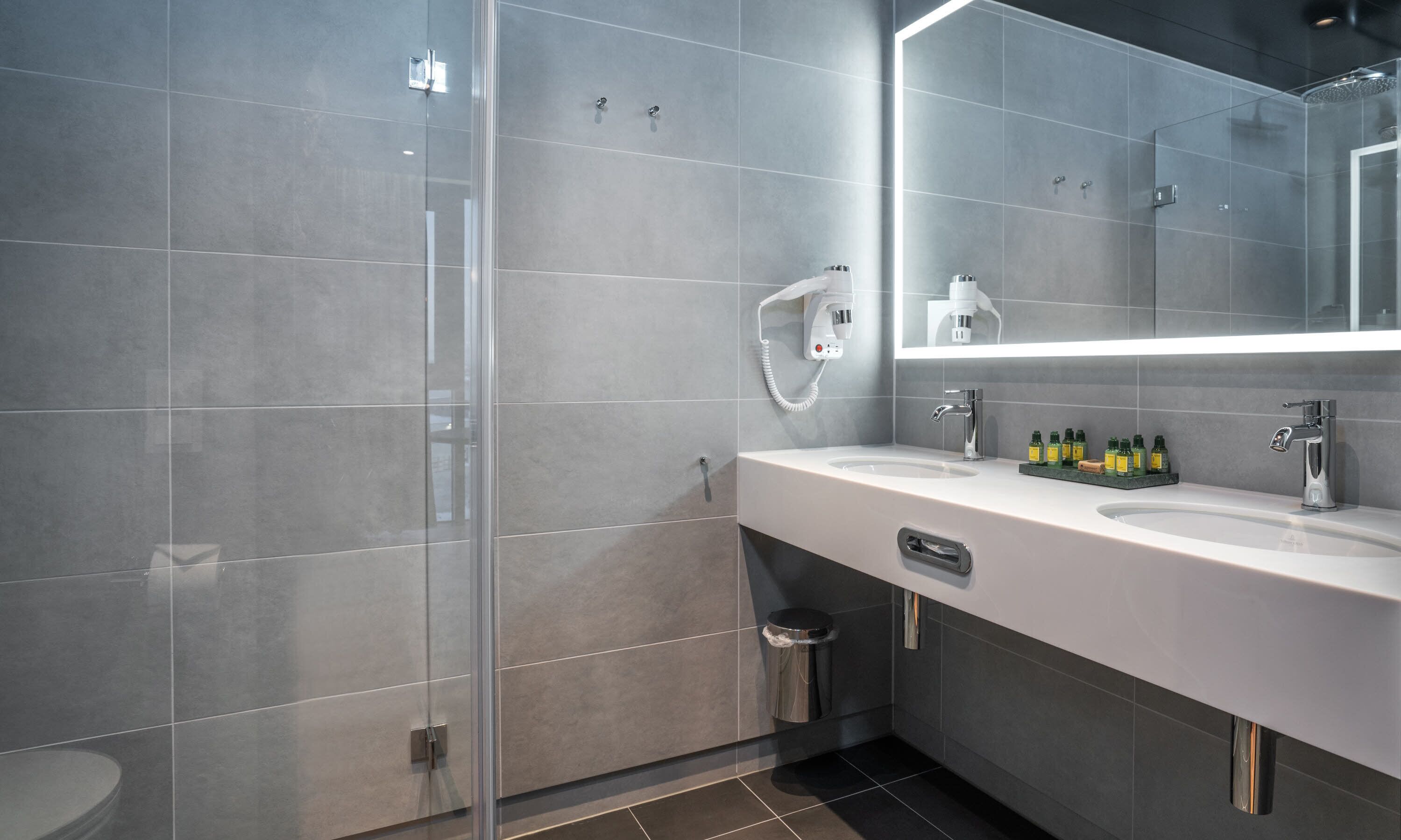 A modern bathroom with a glass shower enclosure, a white sink with a mirror, and a trash bin.