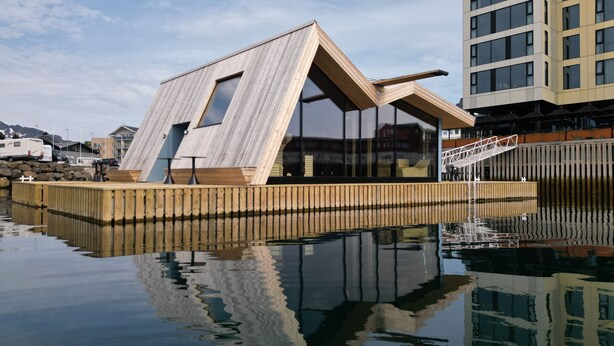 Un edificio moderno con fachada de madera y ventanas amplias, ubicado junto a un cuerpo de agua tranquilo.
