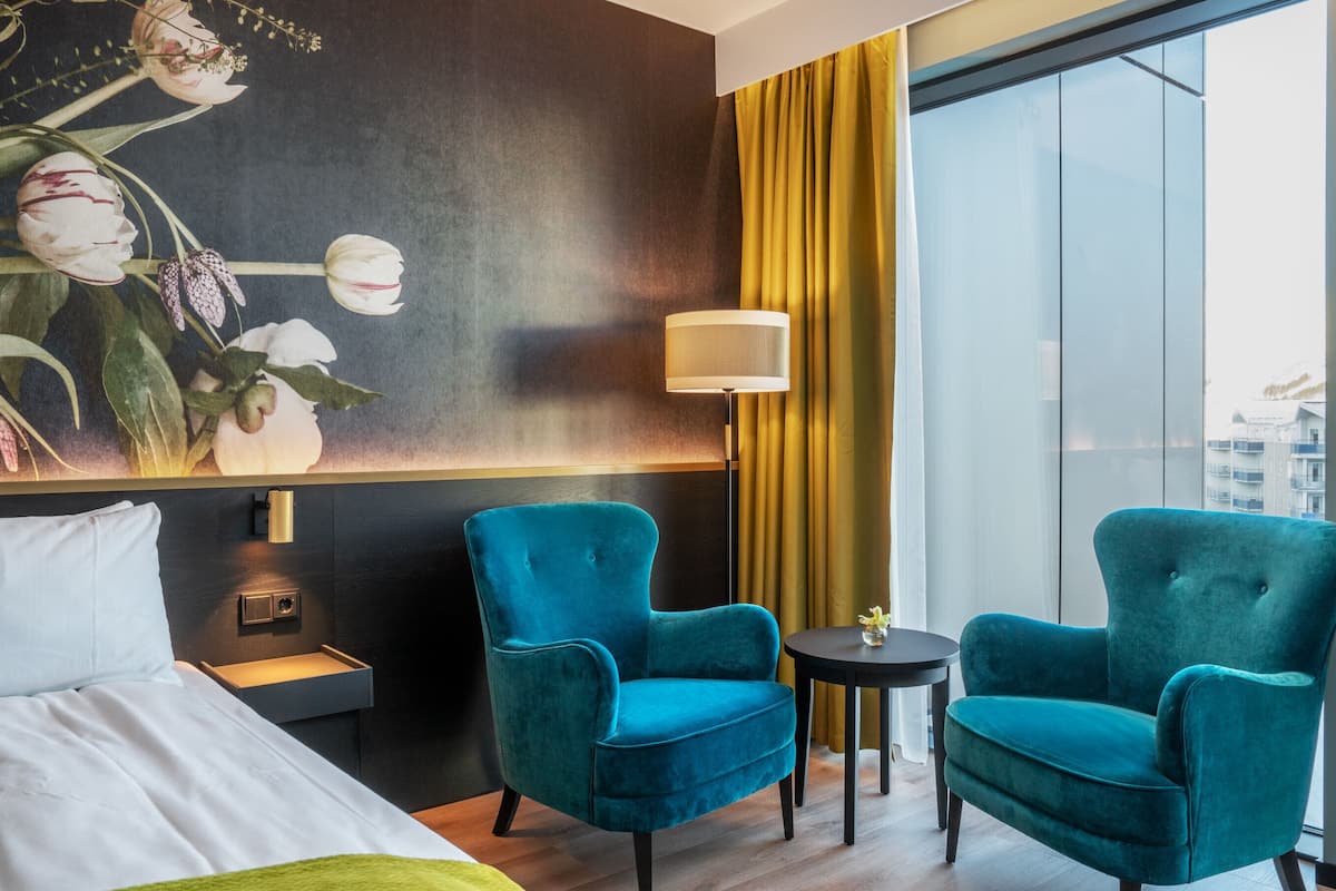 Quarto de hotel com uma cama, duas poltronas verde-água, uma mesa pequena, um abajur e uma janela grande com vista para edifícios.