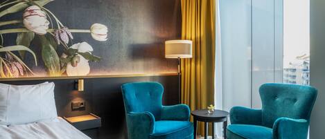 A hotel room with a bed, two teal armchairs, a small table, a lamp, and a large window with a view of buildings.