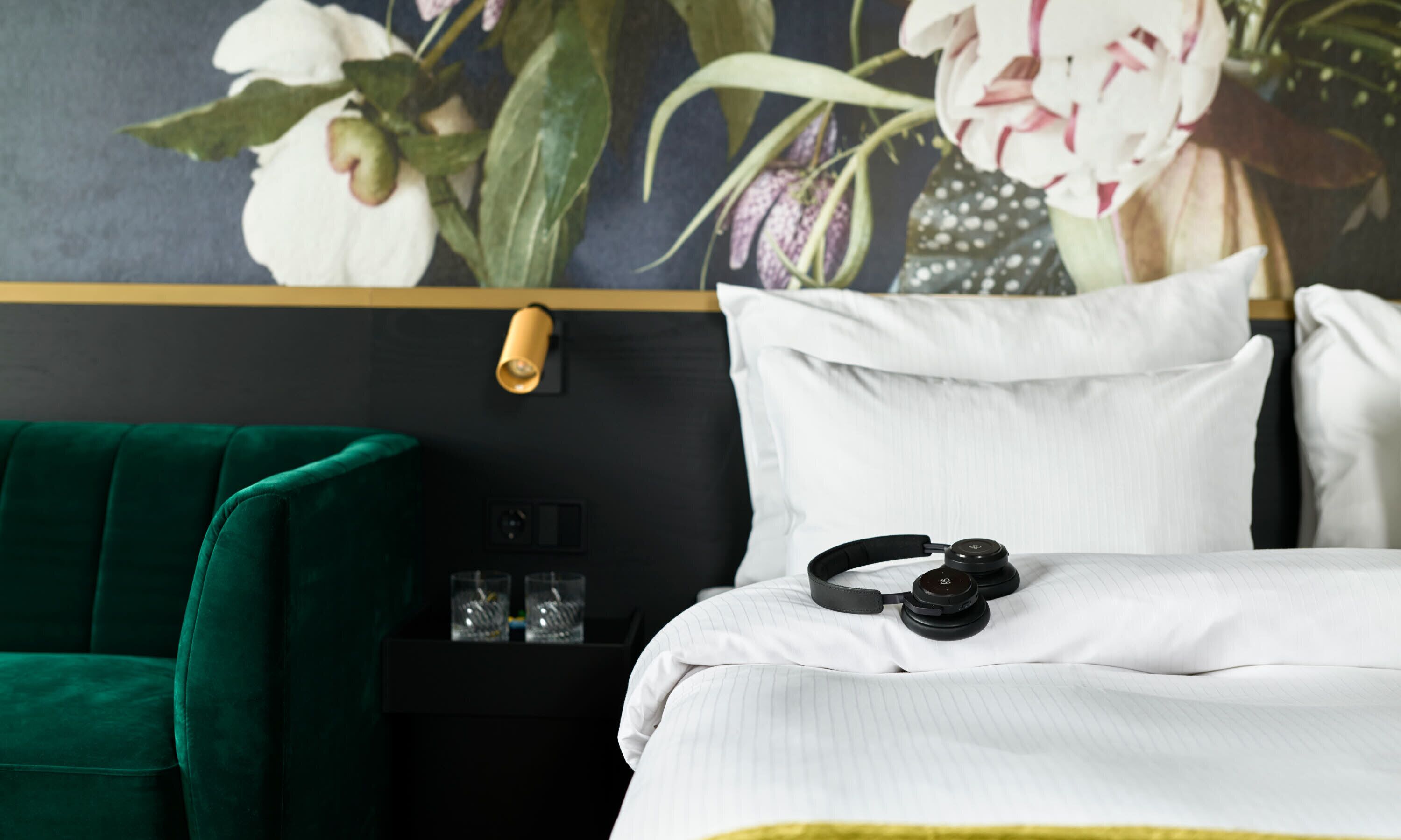 Habitación de hotel con una silla verde, una cama con ropa de cama blanca, un par de auriculares sobre la cama y un estante en la pared con vasos.