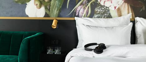 Habitación de hotel con una silla verde, una cama con ropa de cama blanca, un par de auriculares sobre la cama y un estante en la pared con vasos.