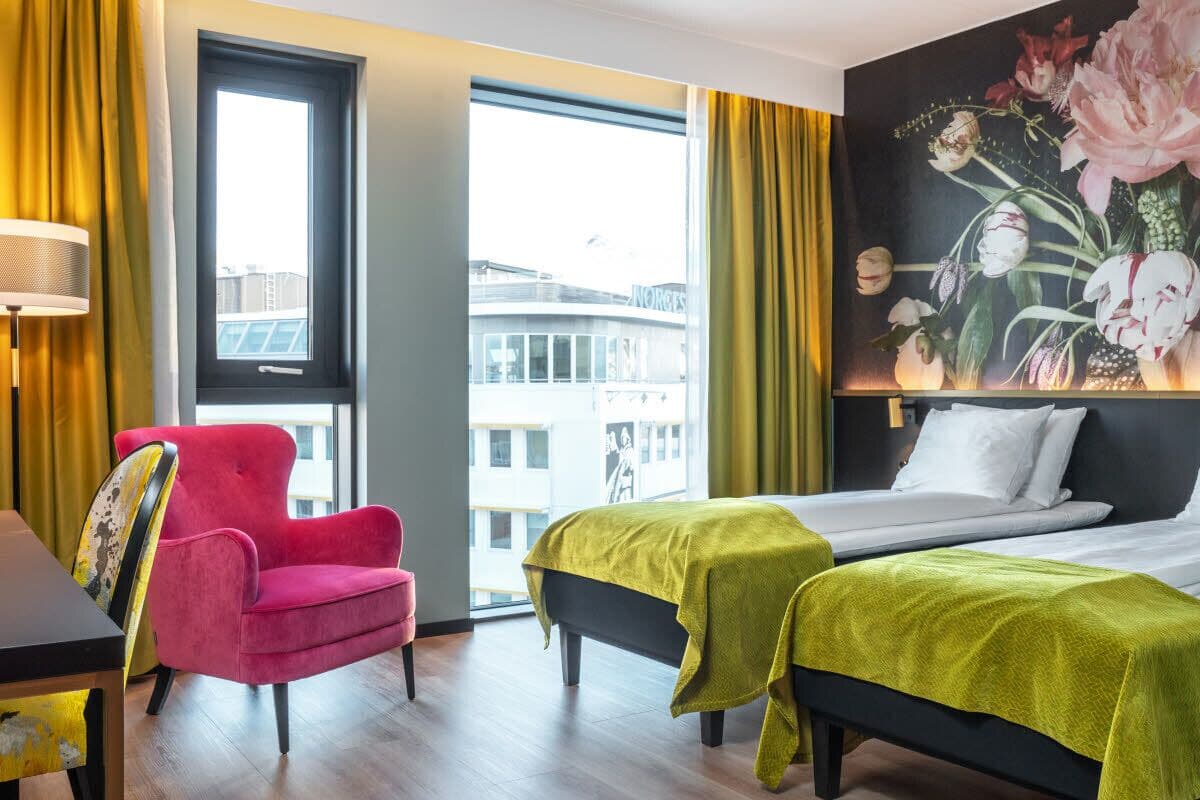 Quarto de hotel com uma cama, duas mesas de cabeceira, uma cadeira, um abajur e um grande quadro com flores na parede.