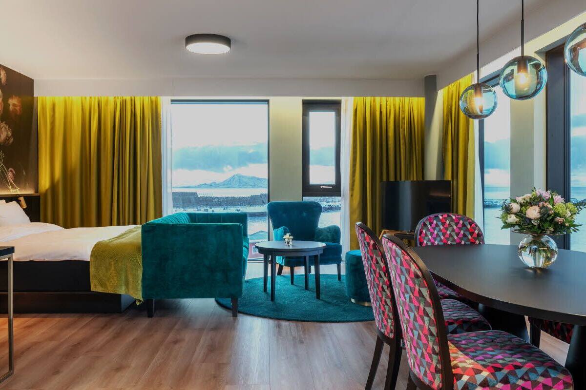 Quarto de hotel moderno com cama, sofá, mesa de jantar e cadeiras. Há uma janela grande com vista para o mar e montanhas.