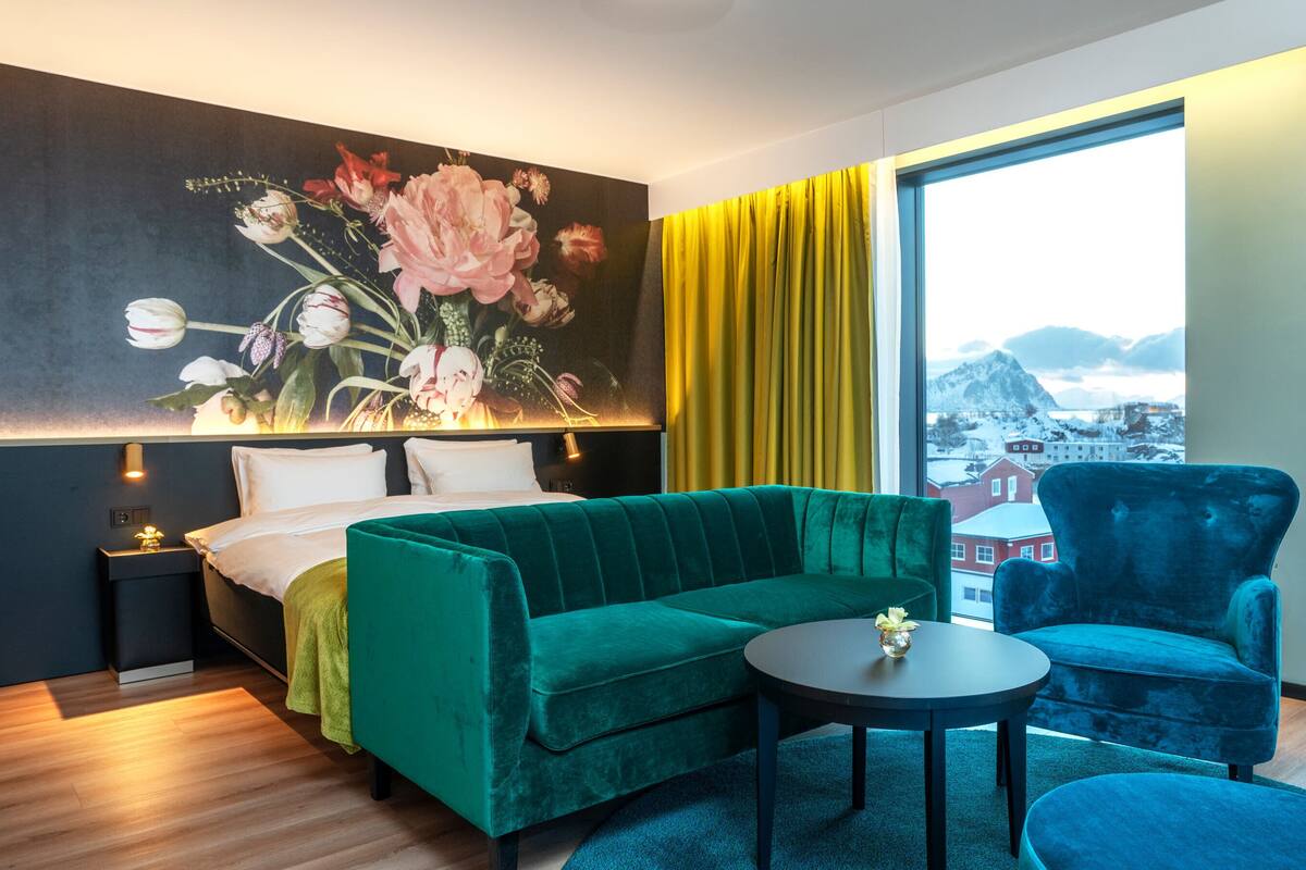Quarto de hotel com uma cama, um sofá verde, um poltrona azul, uma mesinha e uma janela grande com vista para uma paisagem nevada.